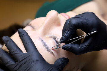 removal of the remnants of unwanted eyebrow hairs of the model after the lamination procedure. the master holds a pair of tweezers in his hands that removes the hairs - obrazy, fototapety, plakaty