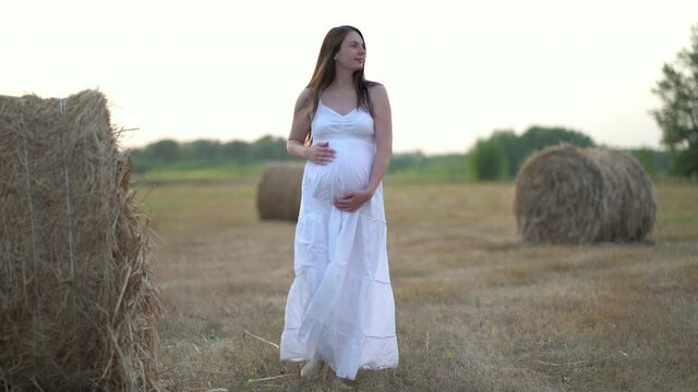 Beautiful pregnant woman in wheat field with haystacks at summer day