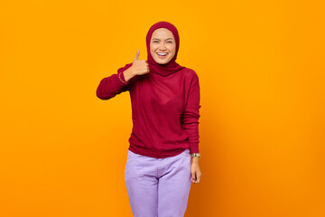 Happy Asian woman showing thumbs up and looking at camera over yellow background