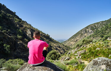 Young caucasian man sitting on a rock looks at the mountainous landscape