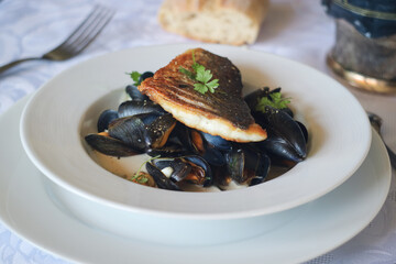 A pan sear crispy filet of sea bass fish on the sea mussels stew in white wine cream sauce in a white plate with parsley leaves on the top for a decoration  with fork and bread in background 