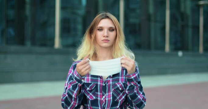 A young beautiful girl in a plaid shirt is standing on the street. The blonde looks at the camera and then puts a white medical mask on her face. She adjusts her mask to make it comfortable.