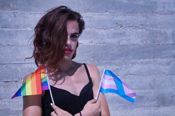 Young Hispanic and transgender girl with her arms crossed and a flag of transsexual pride in her hand and another of gay pride in the other. Concept of transsexuality, inclusion and diversity. - obrazy, fototapety, plakaty