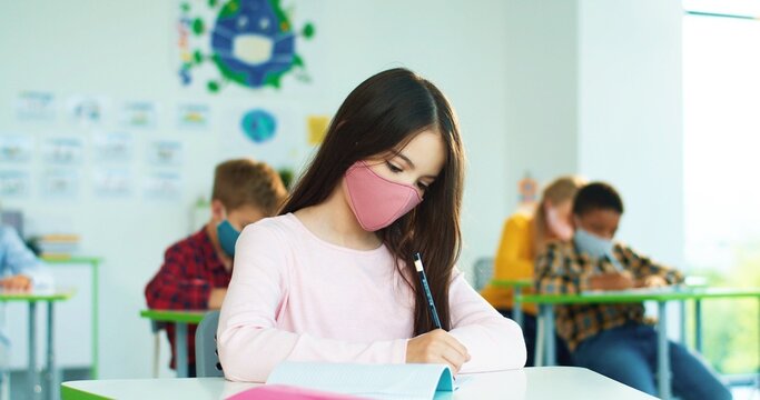 Portrait of beautiful concentrated schoolgirl in face mask studying writing at school while sitting at desk and listening to teacher. - Powered by Adobe