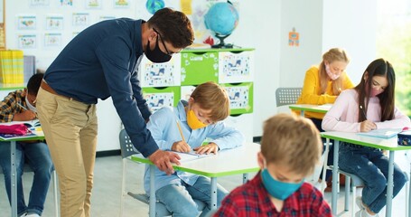 Caucasian young male teacher in mask helping small junior student studying in classroom. Elementary School. Mixed-race children learning after coronavirus lockdown. Lesson, primary school education