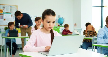 Close up of concentrated Caucasian little girl sitting at desk at school class typing on laptop Children studying in classroom using digital gadgets, computer science class modern elementary education