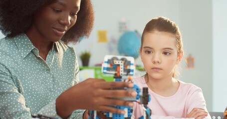 Close up of young beautiful African American woman tutor working in school with Caucasian schoolgirl making toy learning about mechanics. Elementary school lesson, teacher with pupil in science class
