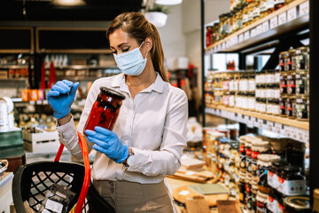 Beautiful young and elegant woman with face protective mask and gloves buying healthy food and drink in a modern supermarket or grocery store. Pandemic or epidemic lifestyle and consumerism concept.