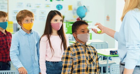 Close up portrait of mixed-race little kids at school in classroom measuring temperature. Female teacher using digital infrared thermometer to measure temperature of pupils, coronavirus concept
