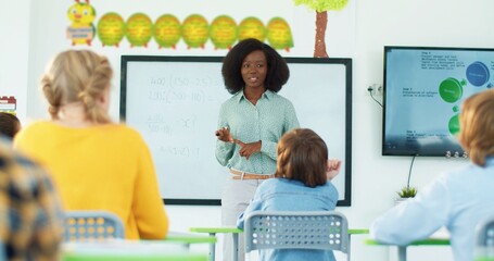 Children study mathematics at elementary school. African American beautiful young teacher standing near the whiteboard explaining lesson to kids at classroom. Education concept, learning