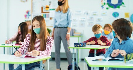 Kids at classroom writing examination test. Caucasian teen schoolgirl in medical mask cheating at exam using cheat sheet while female teacher doesn't see. Post-coronavirus education, pupil concept