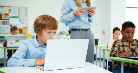 Close up of Caucasian little primary school student sitting at desk in classroom typing on laptop at lesson, female teacher walking behind using tablet. Mixed-race pupils learning at computer class