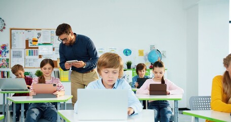 Elementary school computer science class. junior students girls and boys use laptop at lesson. Male teacher explaining and helping schoolgirl at class using tablet Internet technology Modern education