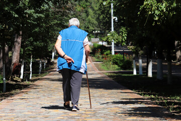 Old woman walking with a cane on a street in town park. Limping person, diseases of the spine, elderly people