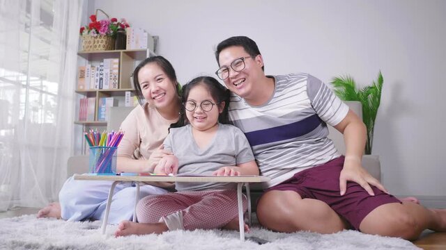 Asian family happy parents and little girl sitting on the floor in living room. Smiling and waving hello goodbye greetings posing in cozy living room, video call