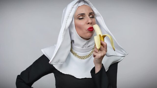 Tempting Caucasian woman in nun costume eating banana smiling looking at camera. Portrait of passionate seductive lady with red lipstick enjoying delicious fruit posing at grey background