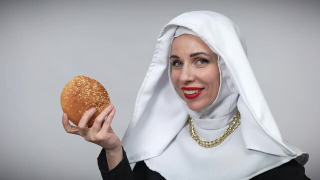 Passionate woman in nun costume licking burger smiling looking at camera. Portrait of sensual seductive Caucasian lady posing at grey background with unhealthy junk food