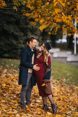 Happy couple walking at the park with yellow leaves on the autumn day. Handsome man and beautiful pregnant woman posing outdoors