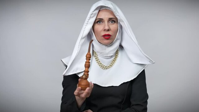Front view of slim woman in nun costume smoking pipe looking at camera. Portrait of beautiful Caucasian lady posing at grey background with tobacco pipe. Sin concept