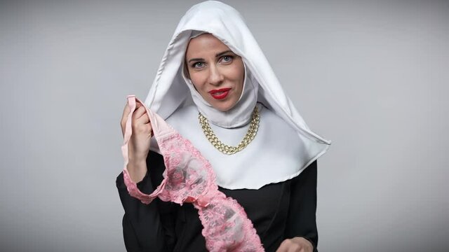 Seductive woman in nun costume holding pink lace bra looking at camera posing at grey background. Portrait of passionate Caucasian gorgeous lady with delicate undergarments smiling