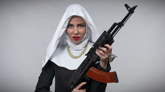 Woman in nun costume holding hunting gun winking looking at camera. Portrait of beautiful Caucasian lady posing with weapon at grey background flirting
