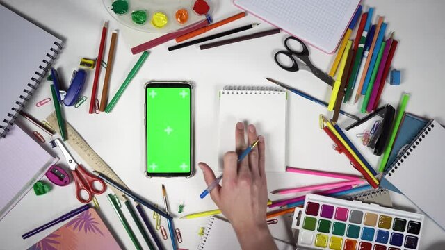 A student holds pen and wants to write information in notebook seen in smartphone with green screen. Mobile with chroma key on display lies on desk among school tools and man swipes content by finger.