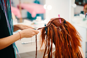 Girl hairdresser weaves woman ginger dreadlocks and cut threads with scissors. Braiding process plait with colored kanekalon. Bright pigtails on head. Hippie style hairstyle. Beauty salon services.