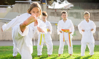 Confident focused preteen girl performing high kick with foot during group taekwondo class on green...