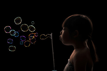 A cute Asian kid girl blowing a lot of colorful bubbles with a black background.
