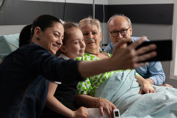 Family taking selfie with sick retired grandmother during medical recovery using modern smartphone in hospital ward supporting her after medicine surgery. Senior woman patient resting in bed