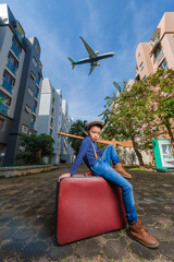 A handsome kid boy about 10years old wearing a vintage dress posing for a photo with an old red vintage suitcase alone with a large plane flying in the background.