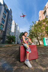 A good looking Asian girl about 8years old wearing a vintage dress posing for a photo with an old red vintage suitcase alone with a large plane flying in the background.