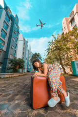 A cute adorable Asian girl about 6years old wearing a vintage dress. An active kid poses for a photo with an old red vintage suitcase alone with a large plane flying in the background.