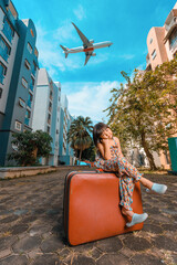 A cute adorable Asian girl about 6years old wearing a vintage dress. An active kid poses for a photo with an old red vintage suitcase alone with a large plane flying in the background.