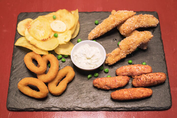 Fast food: onion rings, potatoes and fried chicken, sauce on a dark table, top view 
