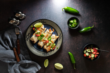Top down view of a dish of taquitos surrounded by ingredients, against a dark background.