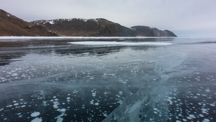 The smooth, shiny surface of a frozen lake. The cracks go deep into the ice. Methane gas bubbles are visible. A mountain range against the sky. Baikal