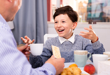 Preteen son talking with father at table with tea during breakfast indoors