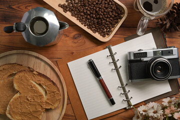 Moka pot, notebook, camera and peanut butter sandwich on wooden table.