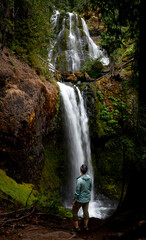 View of a person standing in front of a two-tier waterfall in Oregon. Falls creek falls can be seen with cascading water and green moss in a lush forest. 