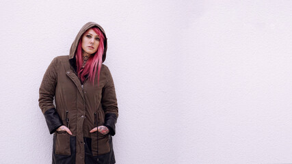 young woman with hooded parka pink hair piercings and tattoos against wall with copy space