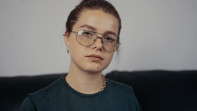 The portrait of a freckled redhead, with her hair caught, wearing her glasses, in slow motion. Dressed in a dark green blouse, sitting on a black sofa, at home. The concept of the woman relaxing at