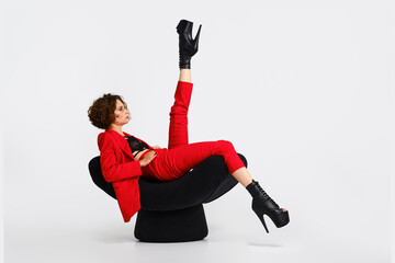 Dancer woman in red pantsui and pole dance boots sits in a soft chair and lift one leg up