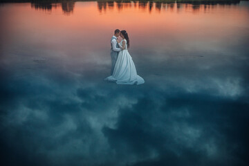 Newlywed couple standing on water at sunset.
