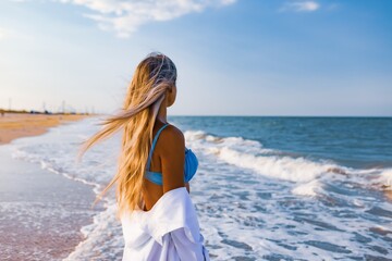 Naklejka premium A slender girl in a gentle blue swimsuit and shirt walks along the sandy beach near the blue sea