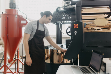 Attractive young man in apron checking roasted coffee beans in cooling tray while using coffee roasting machine