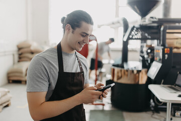 Portrait of handsome modern man wearing apron use phone while standing at coffee roasting machine in local roastery