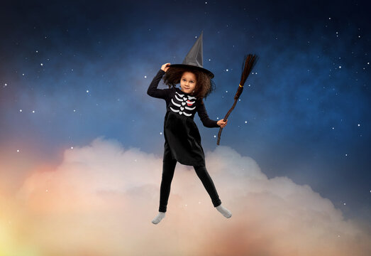 halloween, holiday and childhood concept - smiling african american girl in black costume dress and witch hat with broom over starry night sky and cloud background