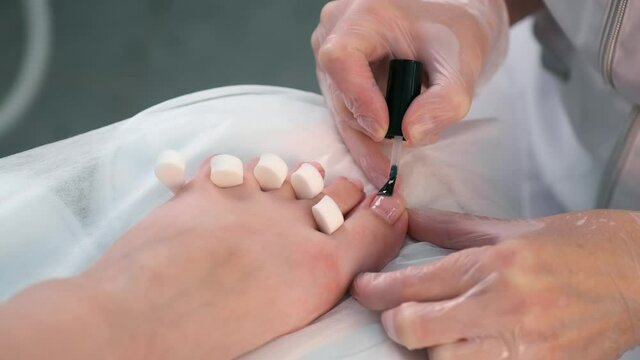 Pedicurist master is applying transparent base coal for shellac on woman's nails on toes, closeup hands. Preparing nails for shellac in beauty salon. Hygiene and care for feet.