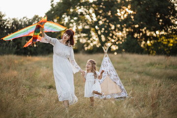 Naklejka na ściany i meble Young pretty mother helping her child to fly a kite, running in the sumer field. Boho woman and her cute little daughter playing with colorful kite together in the meadow. Tee pee tent on background